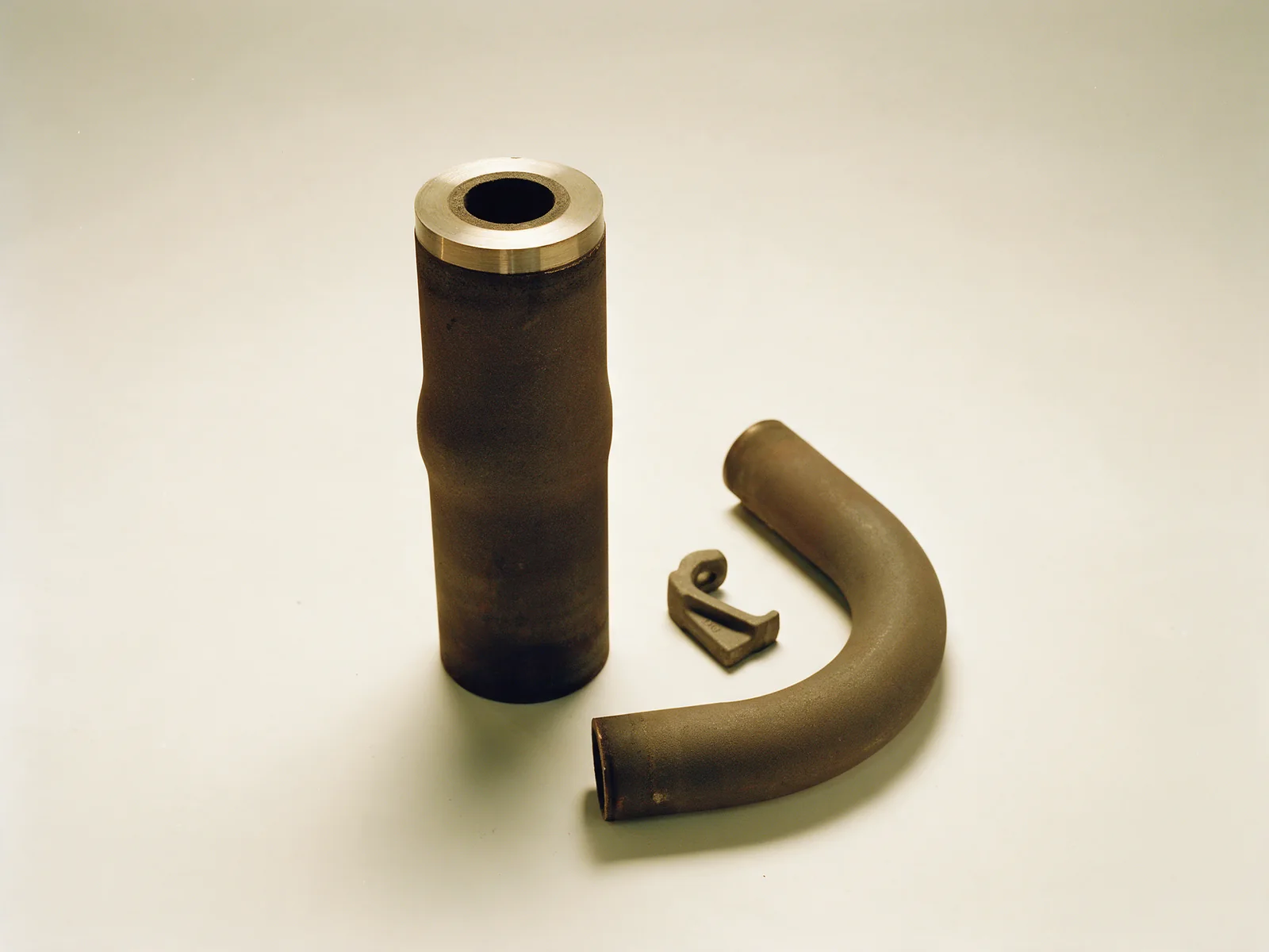 A retired reformer tube section with dark oxide scale and a bright machined cut face, a bent radiant tube elbow and a small cast hanger on an off-white studio surface