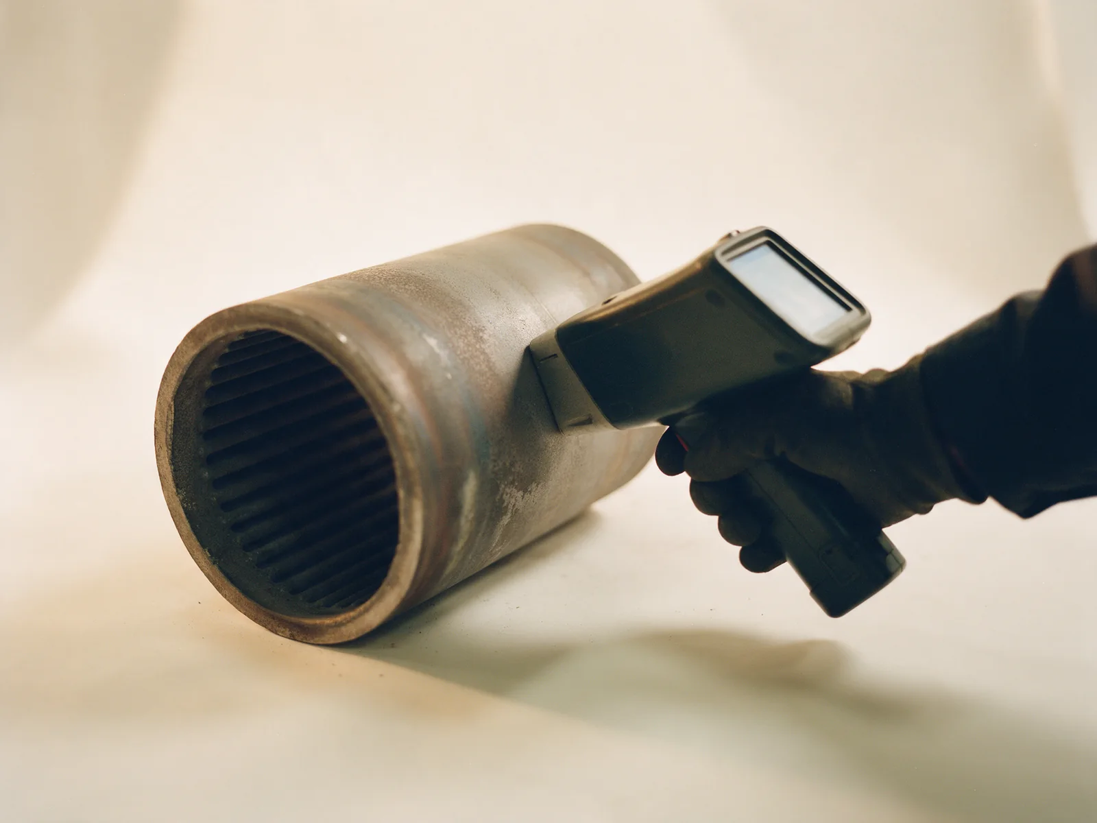A gloved hand pressing a handheld XRF alloy analyzer against the wall of a retired tube section on an off-white studio surface